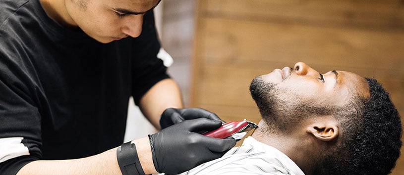 Male barber finishes trimming client's beard with electric clipper.