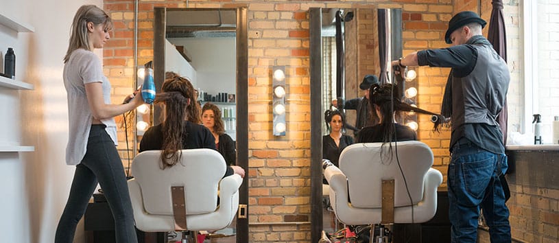 Two hairstylists work in a studio facing mirrors, using various combs and tools to style their client's hair. 